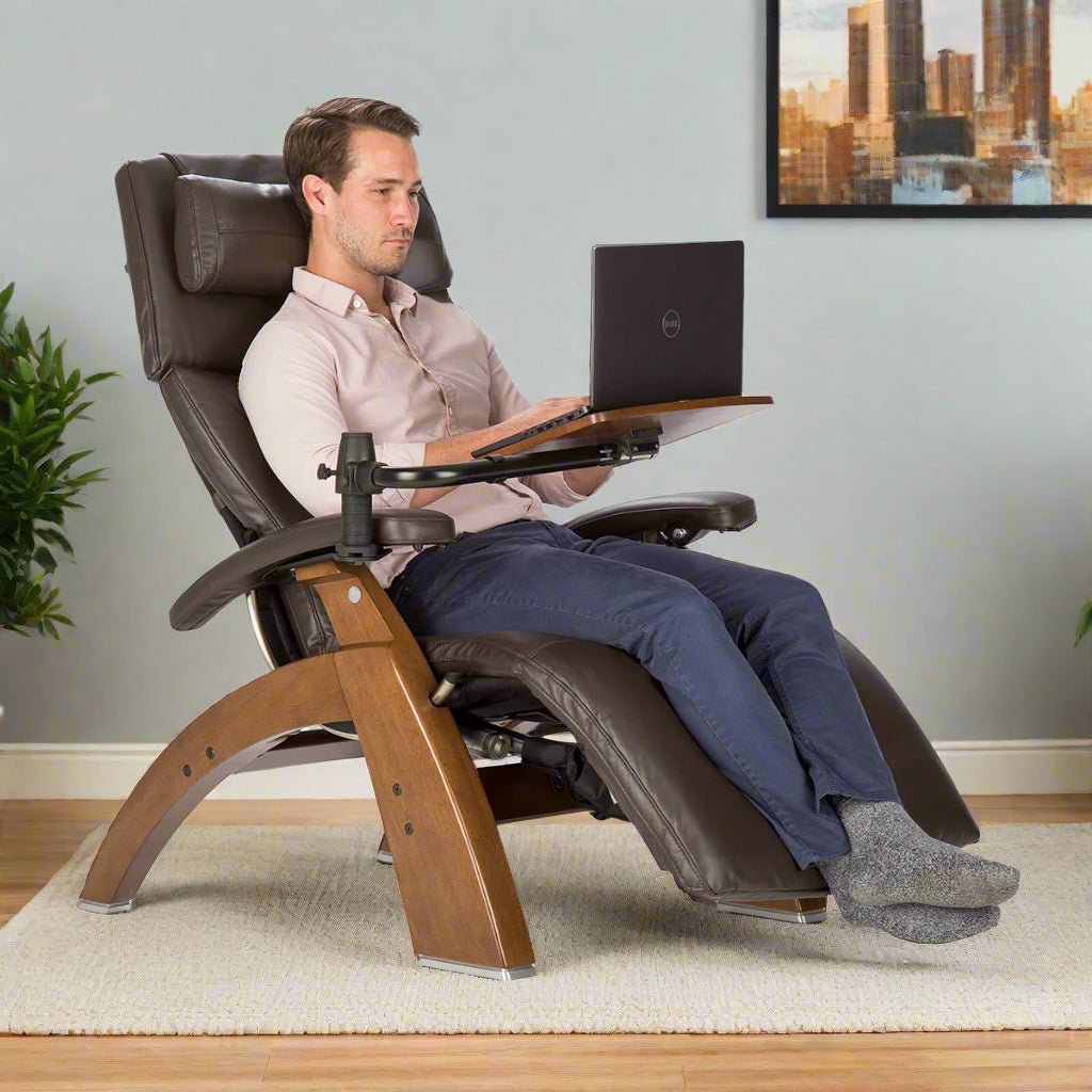 A man works on his laptop while seated in a brown recliner with a built-in desk.
